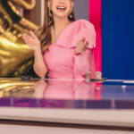 A smiling woman in a pink dress sitting at a news desk, gesturing with her hands.