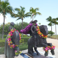 Statue of a man on a horse adorned with colorful leis and palm trees in the background.