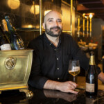 Bearded man enjoys a beer at a cozy bar with bottles and a champagne bucket.