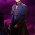 Actor in a dark blue period costume on stage with dramatic purple lighting.
