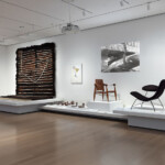 Modern art gallery with sculptures, chairs, and textile art on white platforms.