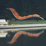 Curved orange lounge chair by a calm pool reflecting its image at dusk.