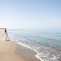 A woman in white dress walking along a calm beach shore.