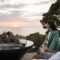 Two people enjoying a sunset by an outdoor fire pit with drinks.