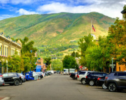 A street lined with parked cars against a backdrop of green mountains under a blue sky.