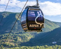 Aspen gondola cable car glides above green mountains under a blue sky.