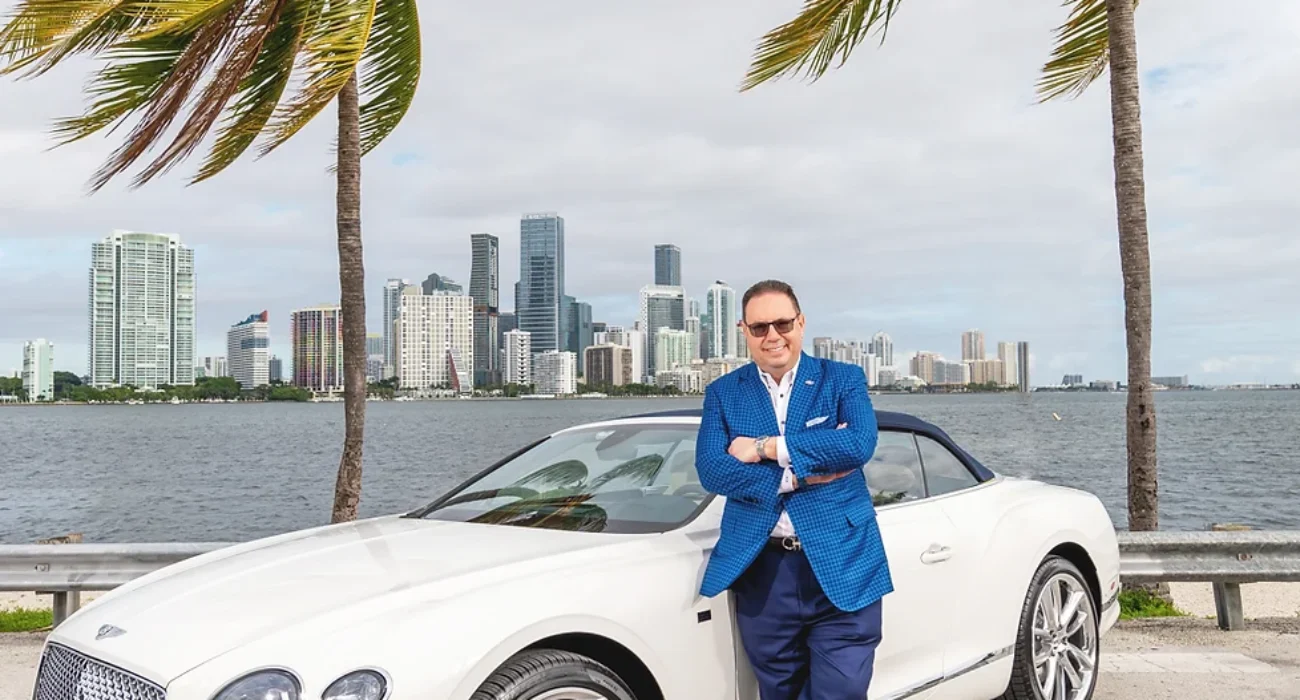 Man in blue suit leaning on a white luxury car by the waterfront with cityscape background.