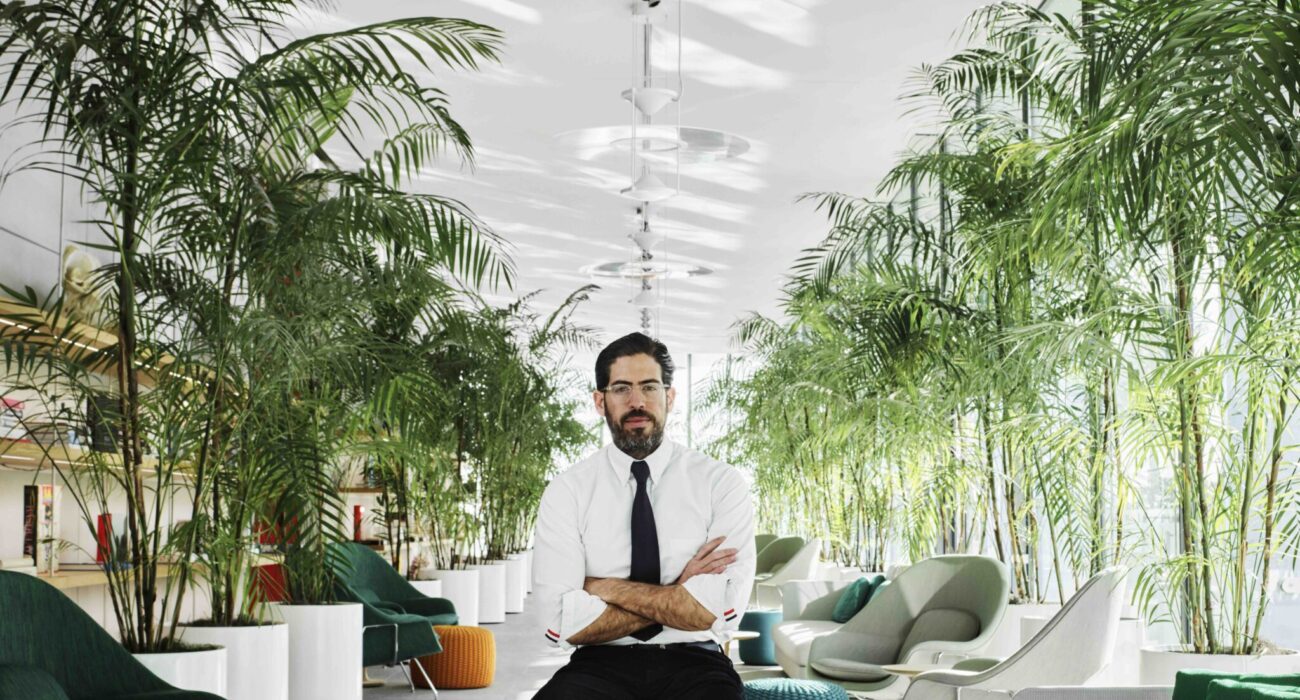 Man sitting confidently on a chair in a modern, plant-filled office space.