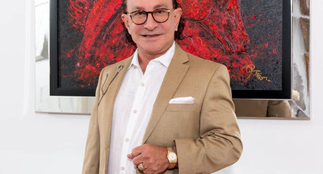 Distinguished man in a beige suit stands confidently indoors against a red artwork backdrop.