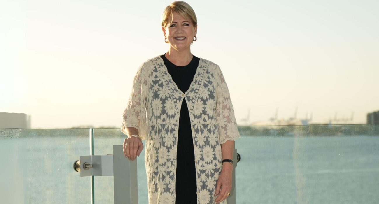 Smiling woman in a black dress and white lace shawl on a balcony at sunset.