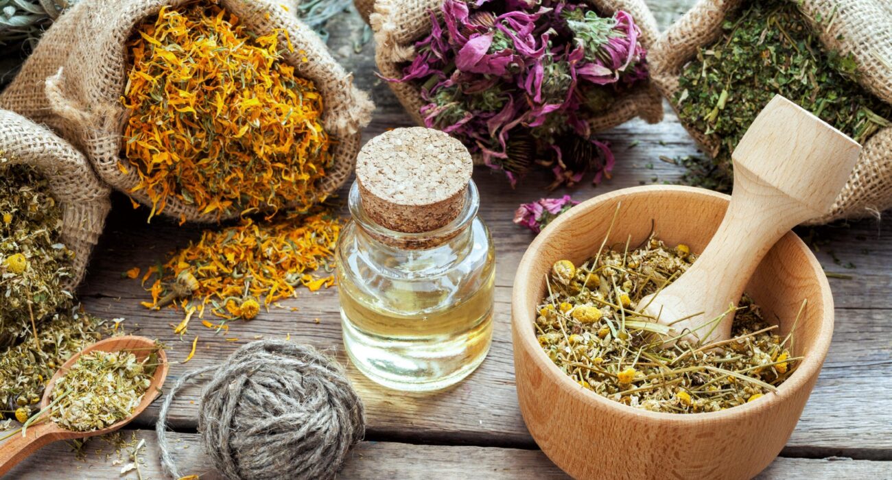 Various dried herbs, essential oil, and a wooden mortar with pestle on a table.