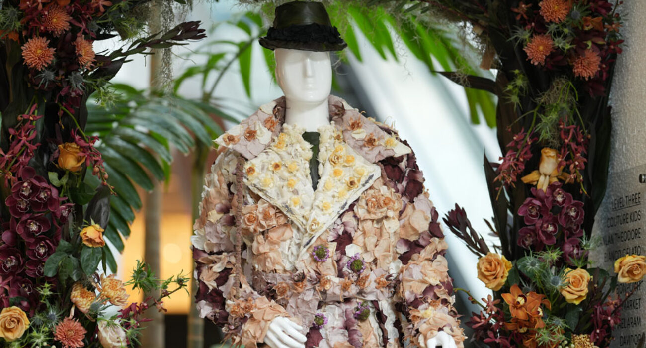 Mannequin dressed in a floral patterned coat and hat among lush greenery.