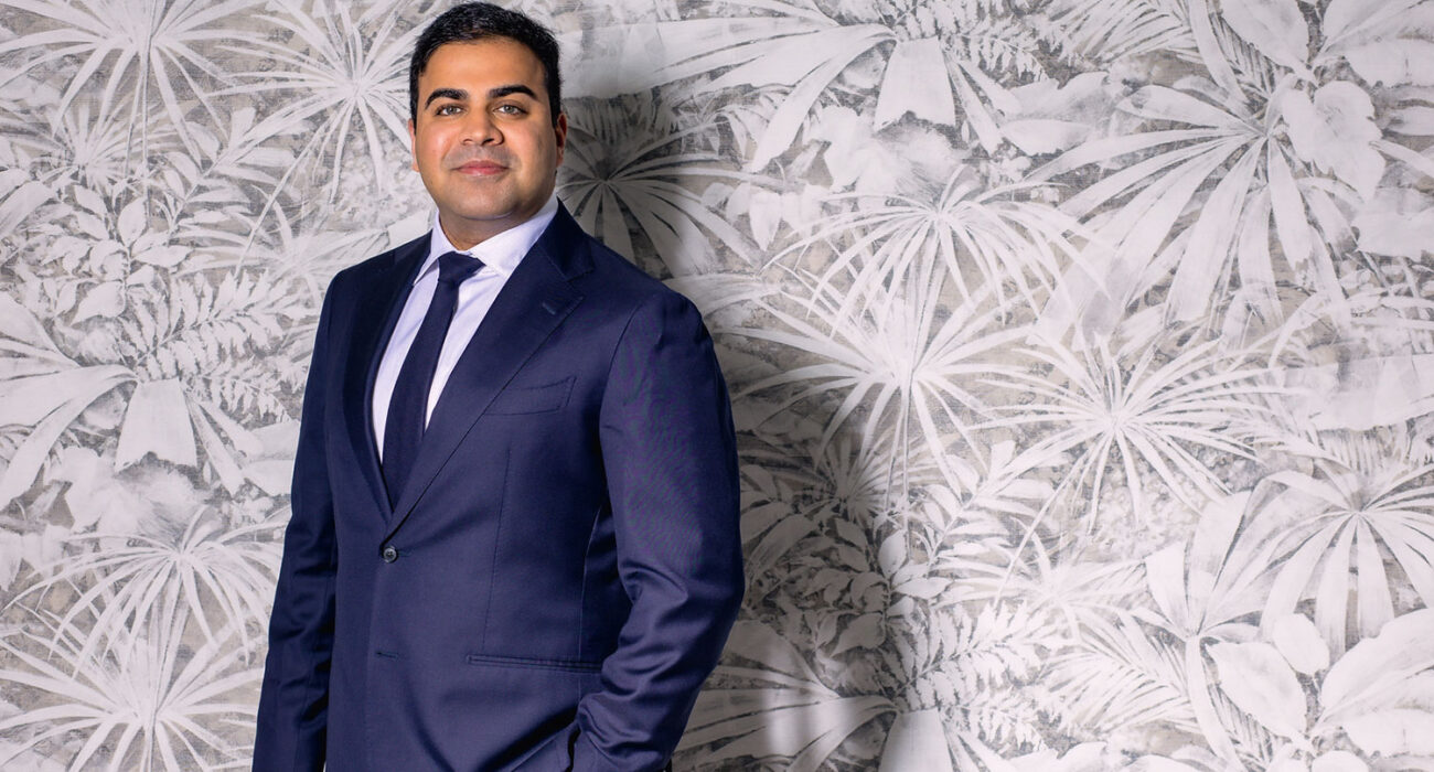 Confident man in a navy suit poses against a textured wall.