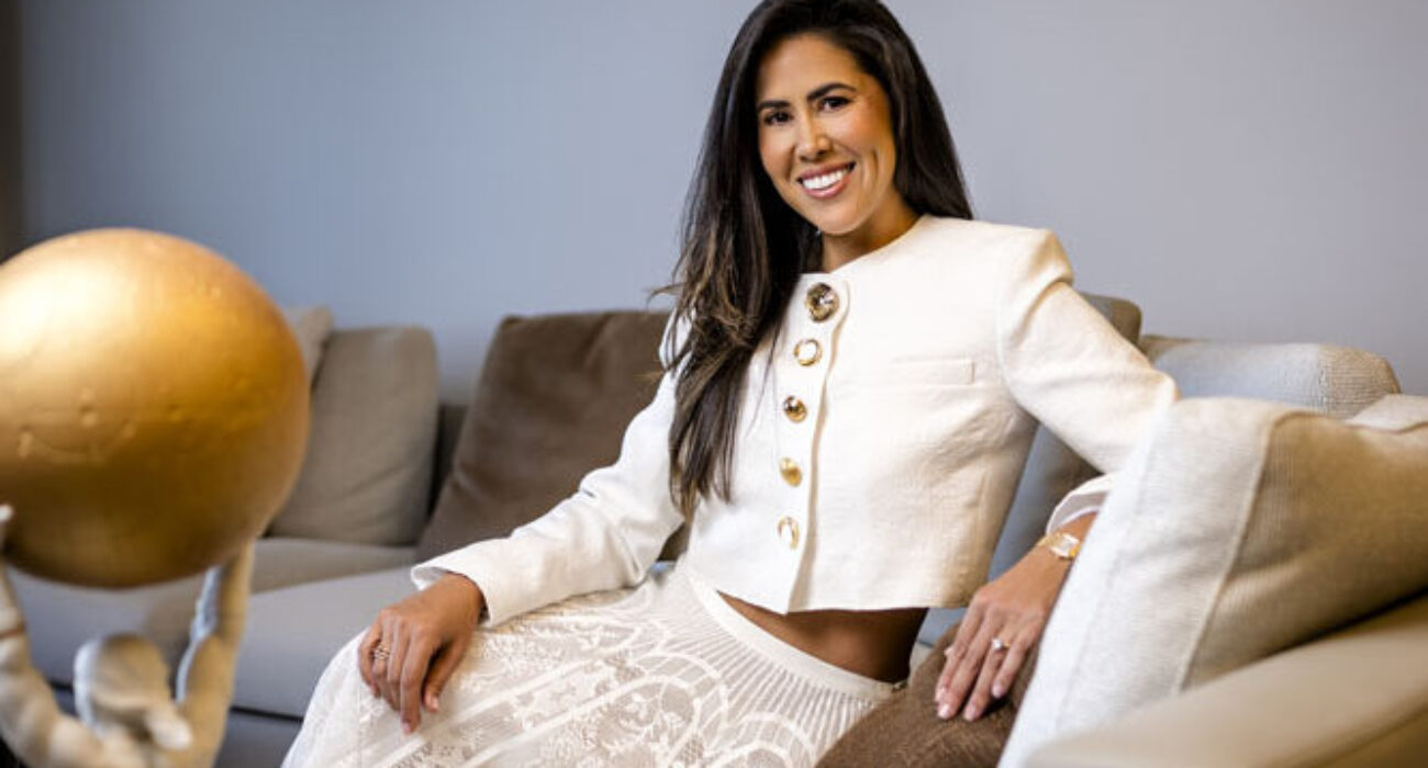 A confident woman in a stylish white outfit sitting on a sofa, smiling warmly.